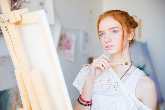 Doordachte aantrekkelijke jonge vrouwenschilder die voor schildersezel met penseel staat en droomt in kunstatelier