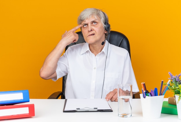 Doordachte blanke vrouwelijke callcenter-operator op een koptelefoon die aan het bureau zit met kantoorhulpmiddelen die de vinger op haar tempel leggen en omhoog kijken