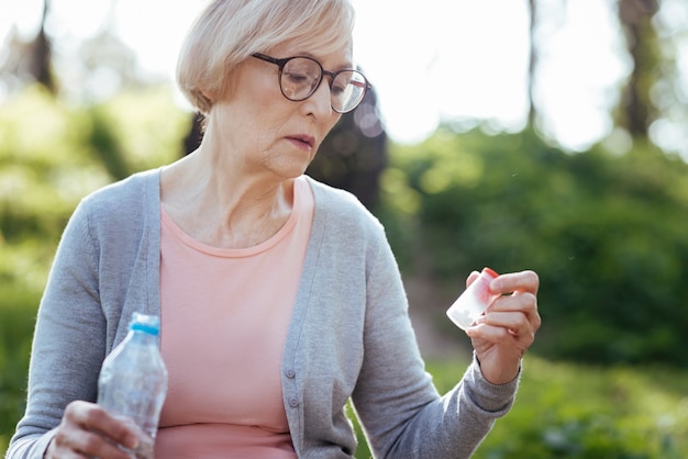 Doordachte boos zieke dame die zich uitgeput voelt en pijnstillers en een fles water vasthoudt terwijl ze buiten zit