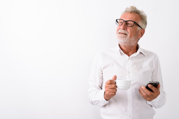 doordachte gelukkig senior bebaarde man glimlachend terwijl koffiekopje en mobiele telefoon met bril op wit