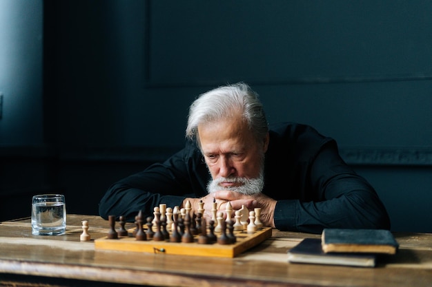 Doordachte grijsharige senior oudere man denken spelstrategie zittend op houten tafel met schaakbord. Medium shot van een peinzende, bebaarde oude man die alleen thuis schaakt.