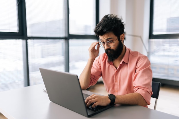 Doordachte Indiase zakenman met vrijetijdskleding die op laptop werkt en aan tafel zit in een lichte kantoorruimte op de achtergrond van het raam