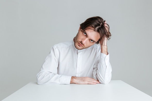 Doordachte jonge brunette man in wit overhemd zittend aan de tafel geïsoleerd op de grijze muur