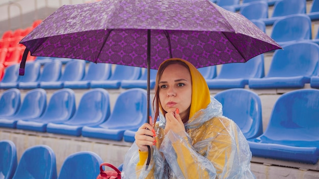 Doordachte jonge vrouw in hoodie en plastic regenjas kin wrijven en wegkijken zittend op de tribune met paraplu in de hand tijdens wedstrijd