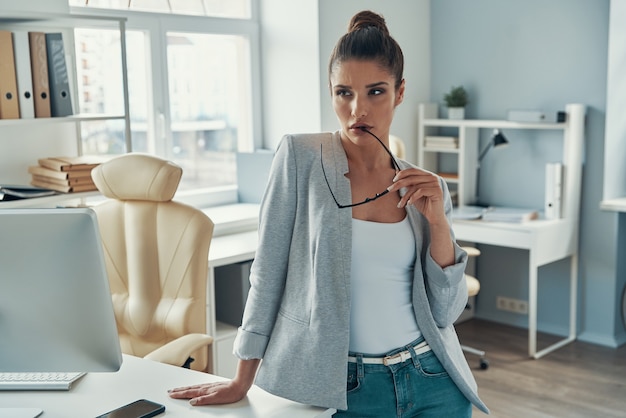 Doordachte jonge vrouw in smart casual die wegkijkt en een bril vasthoudt terwijl ze op kantoor staat