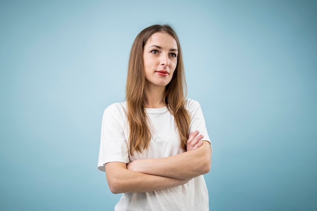 Doordachte jonge vrouw in witte t-shirt met gekruiste armen op blauwe achtergrond