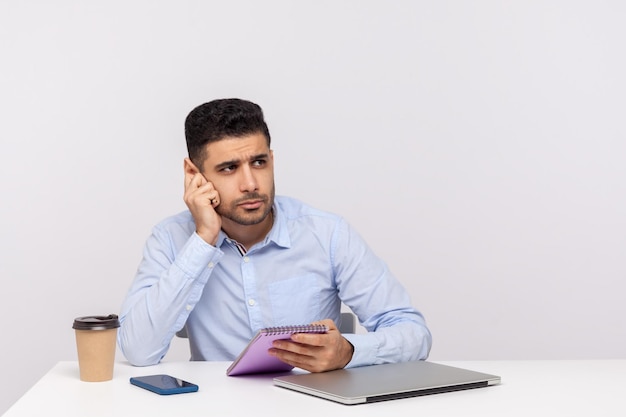 Doordachte journalist of zakenman zittend in kantoor met papieren notitieblok en leunend aan de hand dromen denken over business idee oplossing indoor studio shot geïsoleerd op witte achtergrond