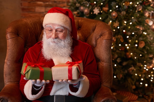 Doordachte kerstman in kostuum zittend in comfortabele fauteuil tegen kerstboom en kiezen...