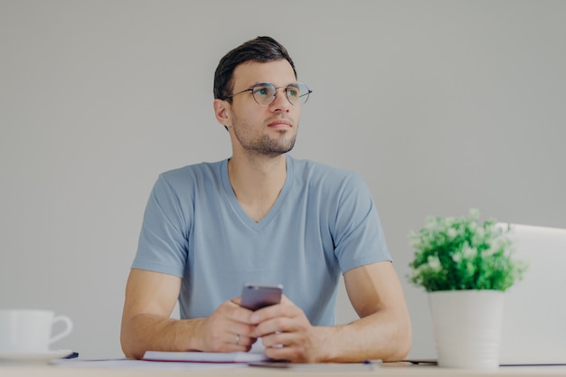 Doordachte mannelijke freelancer werkt op afstand op een laptop, kijkt pensively en houdt moderne mobiele telefoon