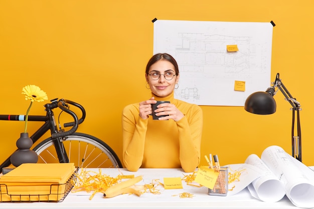 Doordachte professionele vrouwelijke architect drinkt koffie poseert bij dekstop met bouwschets in muur heeft rotzooi op bureau denkt over het creëren van nieuw project draagt bril en coltrui