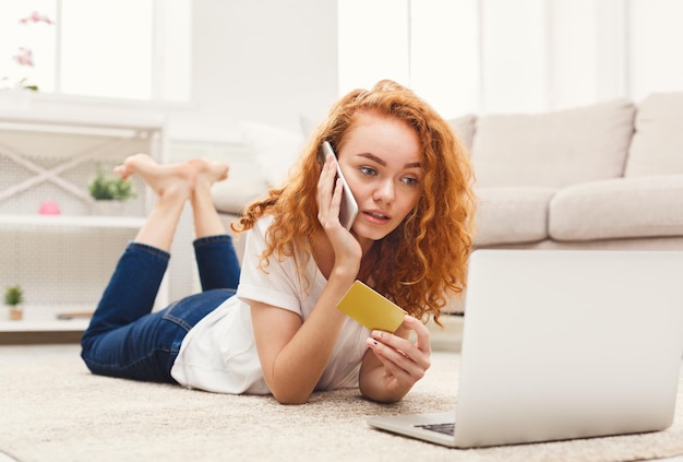 Doordachte roodharige vrouw die online op laptop kiest, met creditcard betaalt en bestelling plaatst op smartphone, liggend op de vloer. Internet winkelconcept
