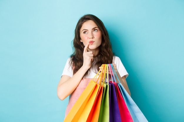 Doordachte vrouw die boodschappentassen vasthoudt, het gezicht aanraakt en wegkijkt terwijl ze nadenkt, een keuze maakt in de winkel, over een blauwe achtergrond staat.
