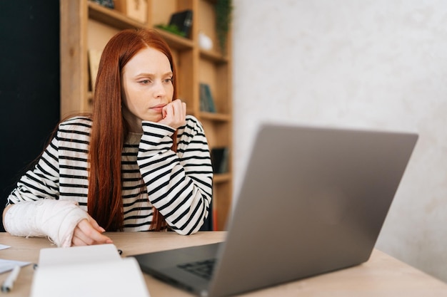 Doordachte vrouwelijke freelancer met gebroken rechterhand gewikkeld in wit gipsverband werkend op laptopcomputer zittend aan tafel met papieren documenten Ongezonde zakenvrouw die op afstand vanuit huis werkt