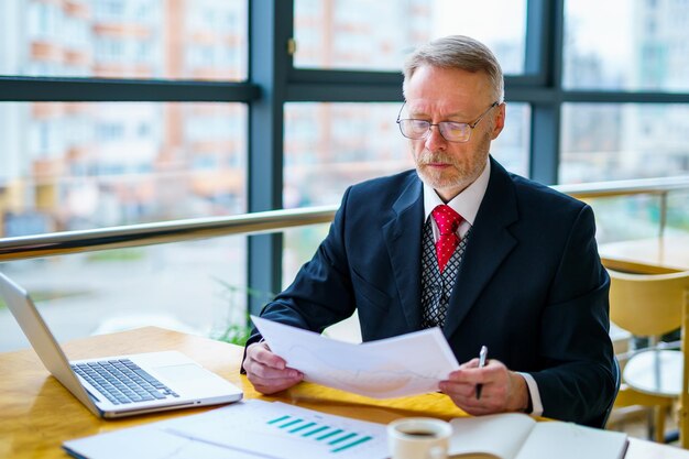 Doordachte zakenman van middelbare leeftijd in pak met een laptop op tafel terwijl hij met documenten werkt Rode stropdas en donker pak