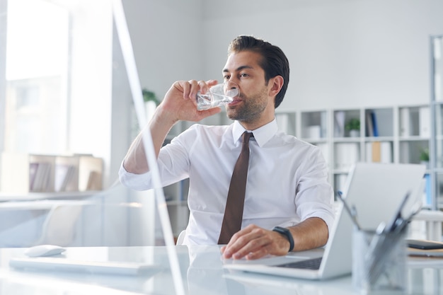 Dorstige jonge makelaar met glas water zittend achter computermonitor in kantoor