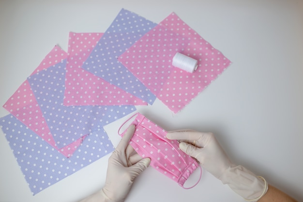 Dotted cloth for face mask sewing on white table. How to sew protective mask, woman hands in white gloves hold handmade cloth mask.