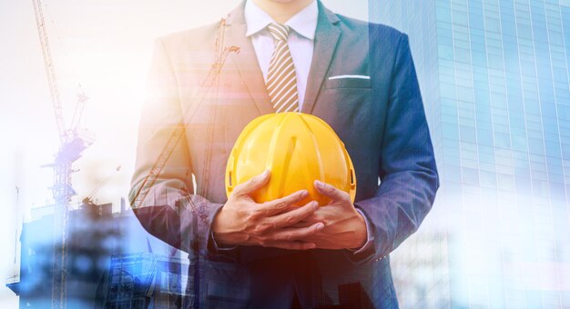 Double Exposure of building under construction and man engineer is holding helmet wearing a suit thinking of ways and ideas for building new buildings in the future.