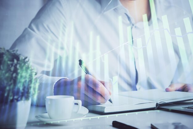 Double exposure of man's hands writing notes of stock market with forex chart