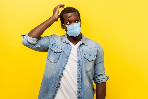 Doubt, need to think. Portrait of uncertain young man with surgical medical mask looking up, scratching head while thinking with confused puzzled face. indoor studio shot isolated on yellow background