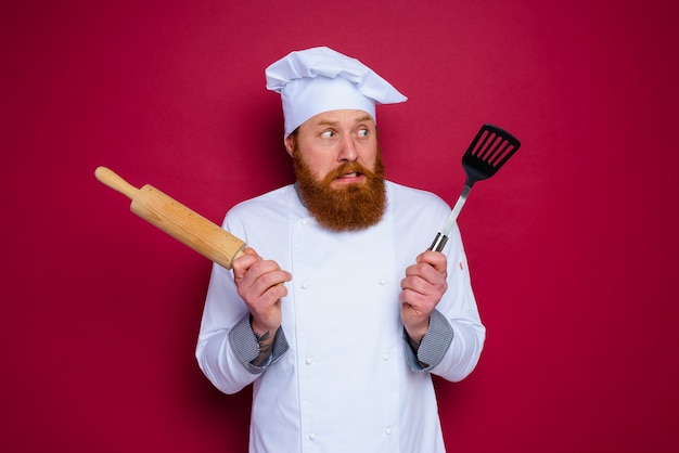 あごひげと赤いエプロンのシェフと疑わしいシェフは、木製の麺棒を保持します