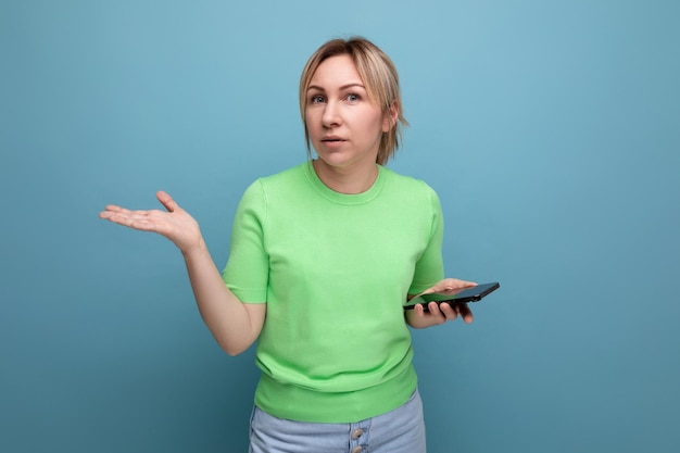 Doubtful attractive blond girl holding a smartphone in her hand on a blue background with copy space