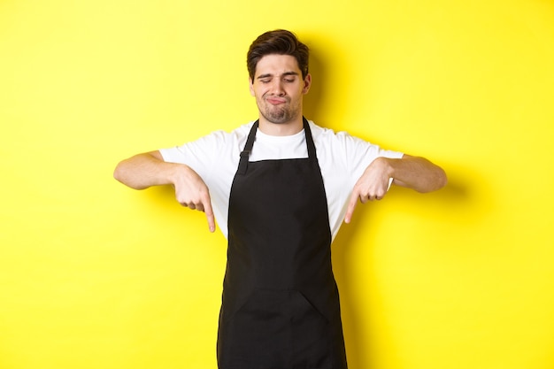 Doubtful barista complaining, pointing fingers down and grimacing displeased, standing in black apron against yellow background