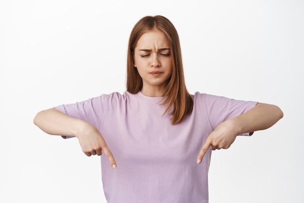 Doubtful blond woman, furrows and points down suspicious, express disbelief, upset about smth, disagree or dislike advertisement, standing against white wall.