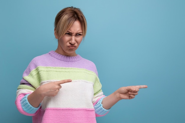 Photo doubtful blond young woman in a striped sweater shows her hand to the side and makes a face on a