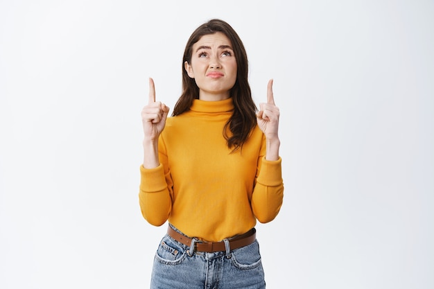 Doubtful and disappointed brunette woman grimacing, pointing and looking up at something bad, feeling uncomfortable or upset, standing on white wall