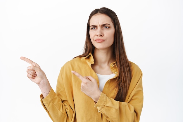 Cliente femminile dubbioso e dispiaciuto che punta guardando a sinistra con accigliata faccia delusa non piace qualcosa che ha dubbi essendo scettico sul prodotto sfondo bianco