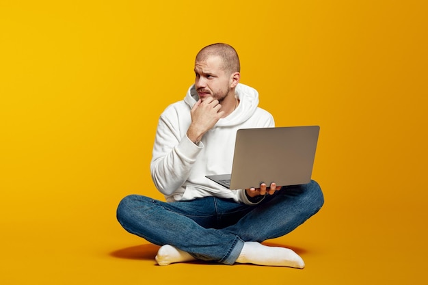 Doubtful guy sitting on floor and holding laptop while looking away isolated over yellow background