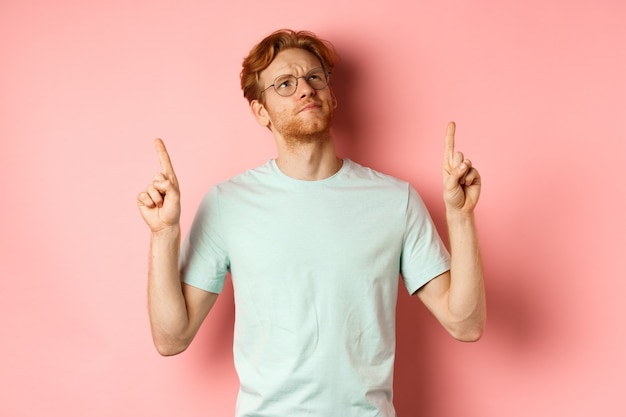 Doubtful redhead guy in glasses frowning looking and pointing fingers up at something disappointed s...