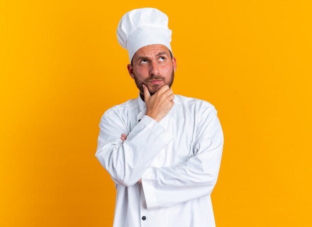 Doubtful young caucasian male cook in chef uniform and cap keeping hand on chin looking up isolated on orange wall with copy space