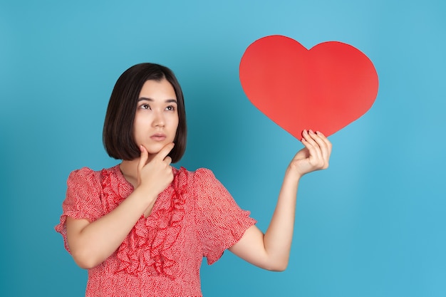 doubting Asian woman holds a large red paper heart in her hands and rubs her chin with her hand