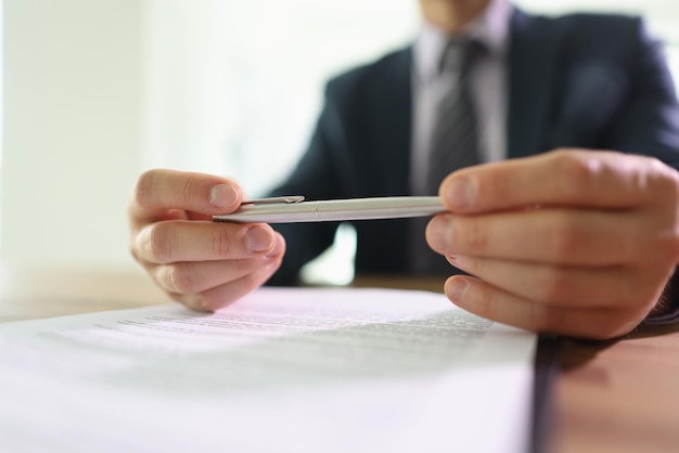 Doubting businessman holds ball pen in hands above contract papers sitting at table in office