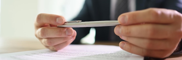 Doubting businessman holds ball pen in hands above contract papers sitting at table in office