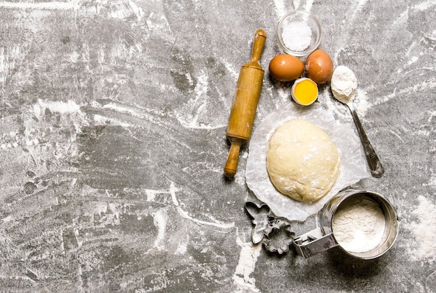 The dough and the ingredients - eggs, flour and tools - a rolling pin and shape. On the stone table. Free space for text . Top view