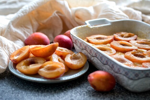 Dough with halves of fresh plums in a ceramic mold and sliced plums on a blue plate