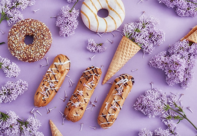 Doughnuts and cream cakes lie on the table among lilac flowers on purple paper. spring and sweets, delicious cakes. still life and summer mood, top view. donuts