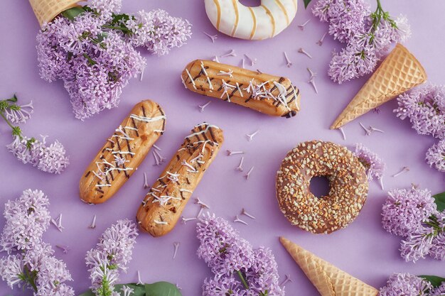 Doughnuts and cream cakes lie on the table among lilac flowers on purple paper