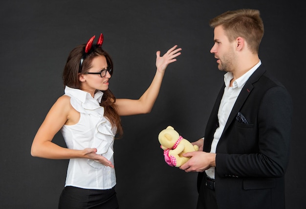 Down with love. Devil woman angry at man with Valentines bear. Corporate Halloween party. Workplace conflict. Be wicked for Halloween.