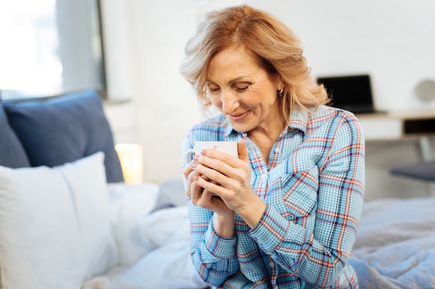 Draag beker. Vrolijke positieve volwassen vrouw die haar dag begint met een kop warme drank terwijl ze in de slaapkamer zit