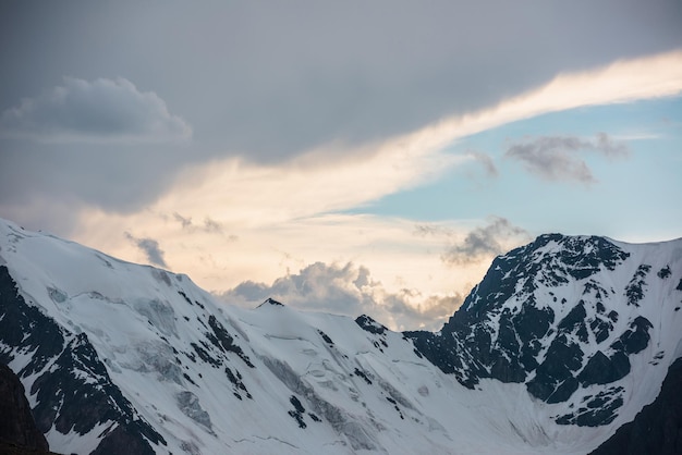 Dramatic aerial landscape with large snowy mountain range under cloudy sky in golden sunset sunlight Awesome alpine scenery with snow mountains under gold clouds in blue sky at changeable weather