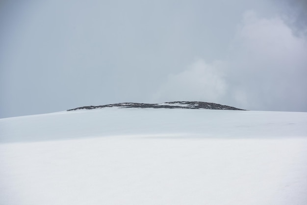 Dramatic alpine landscape with huge snowy mountain dome with rocky top in sunlight under gray cloudy sky Awesome mountain scenery with high snow mountain in dome shape in center at changeable weather