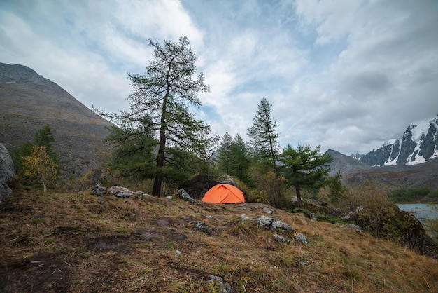 Photo dramatic landscape with alone orange tent on forest hill among rocks and autumn flora with view to large snowy mountain range under cloudy sky lonely tent and fading autumn colors in high mountains