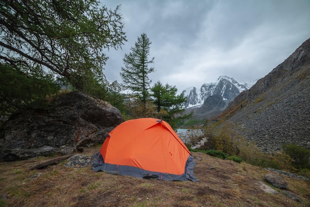 Photo dramatic landscape with alone orange tent on forest hill among rocks and autumn flora with view to large snowy mountain range under cloudy sky lonely tent and fading autumn colors in high mountains