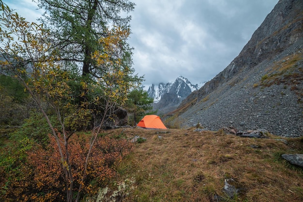 Dramatic landscape with alone orange tent on forest hill among rocks and autumn flora with view to large snowy mountain range under cloudy sky Lonely tent and fading autumn colors in high mountains