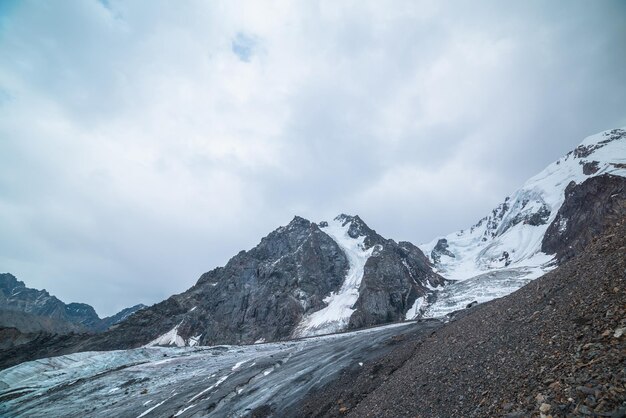 写真 氷河の舌と氷の落下の劇的な風景 大きな雪山脈に 灰色のの空の下でい岩の頂点 長い氷河の高さ 暗い風景の山々