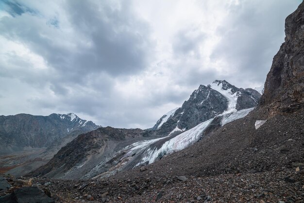 写真 灰色の曇り空の下に鋭い岩の尖塔を持つ大きな雪山を背景に氷河が垂れ下がった劇的な風景高地に氷の雪庇を持つ氷河曇りの山の暗い風景