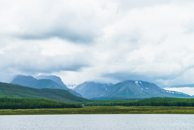 Dramatic landscape with mountain lake and forest on hills in sunlight and snowy mountains in low clouds in changeable weather. High mountains in cloudy sky and lake near green grasses and forest hills